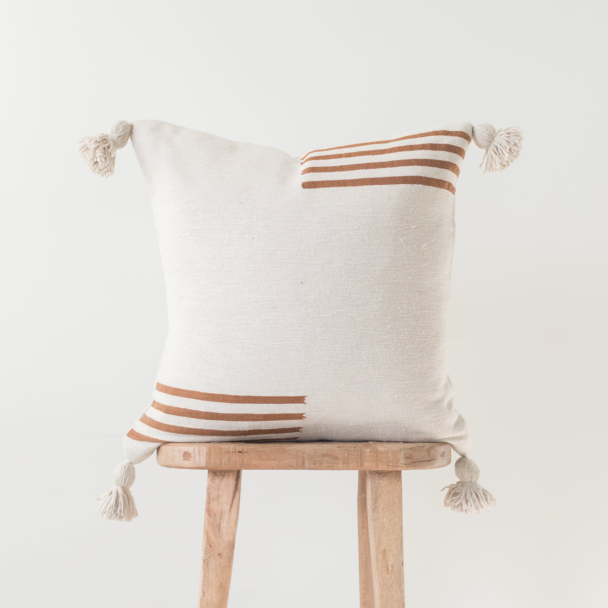 Cream linen pillow with rust and tan stripes and corner tassels, displayed on wooden stool.