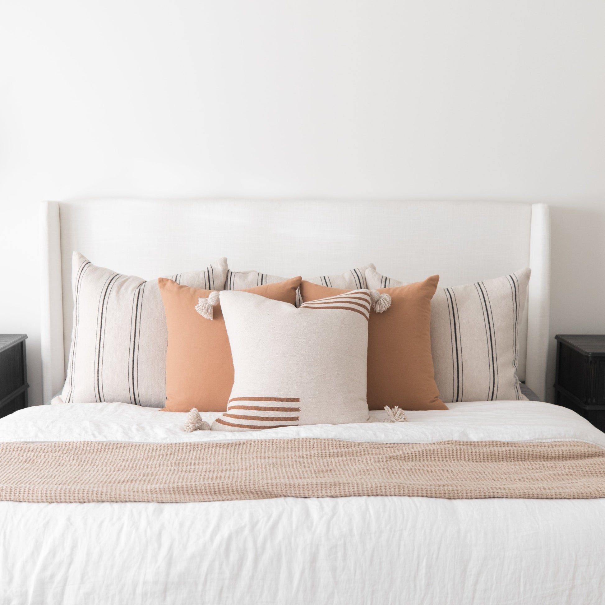White upholstered bed headboard with striped and solid decorative pillows in neutral tones and a textured throw blanket.