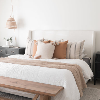 Woven pendant lamp with natural fiber shade hanging above a styled bedroom with white bedding and decorative pillows.