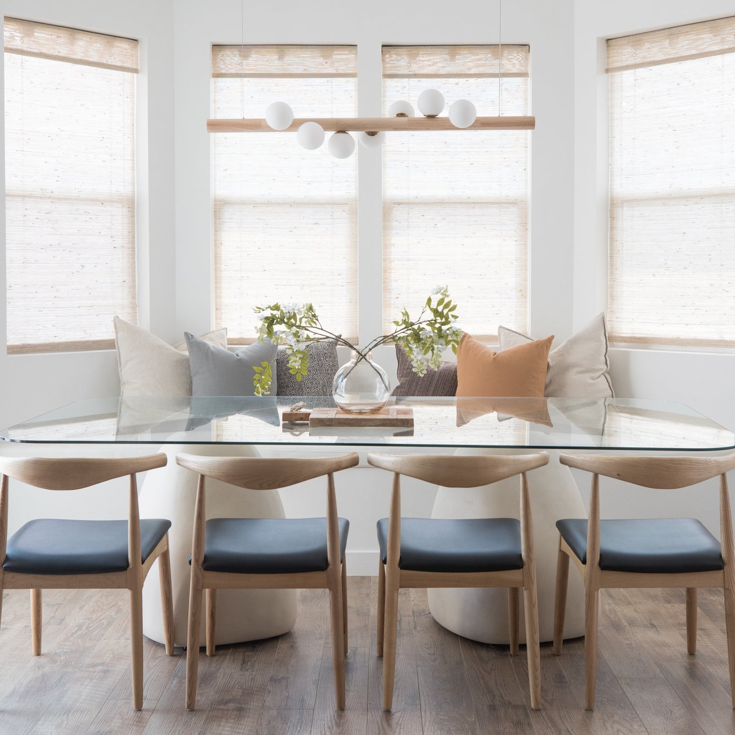 Modern dining room with white table, four wooden chairs with blue seats, and natural linen Roman shades on windows.