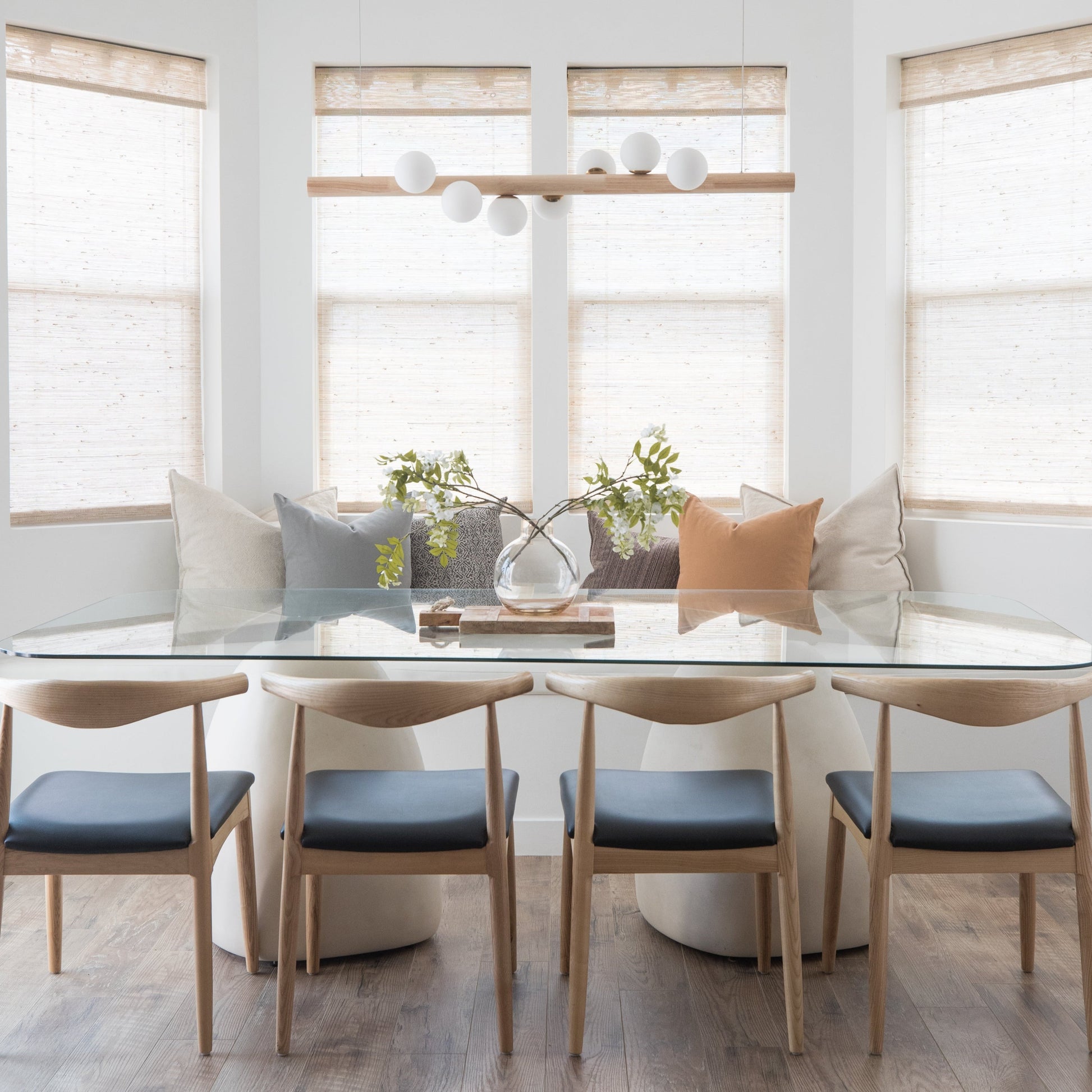 Modern dining room with white table, four wooden chairs with blue seats, and natural linen Roman shades on windows.