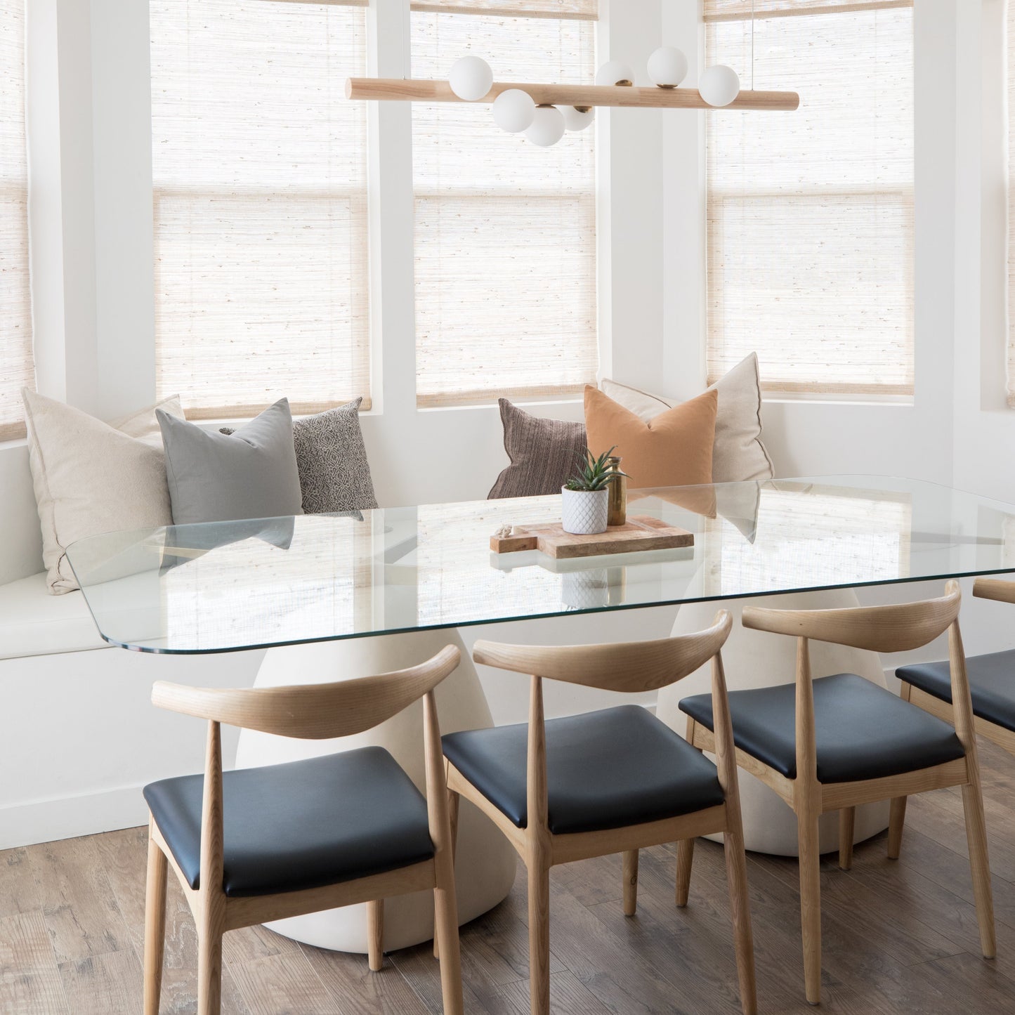 Dining table with glass top and three wooden chairs with black seats in a bright room with windows and cushioned seating.