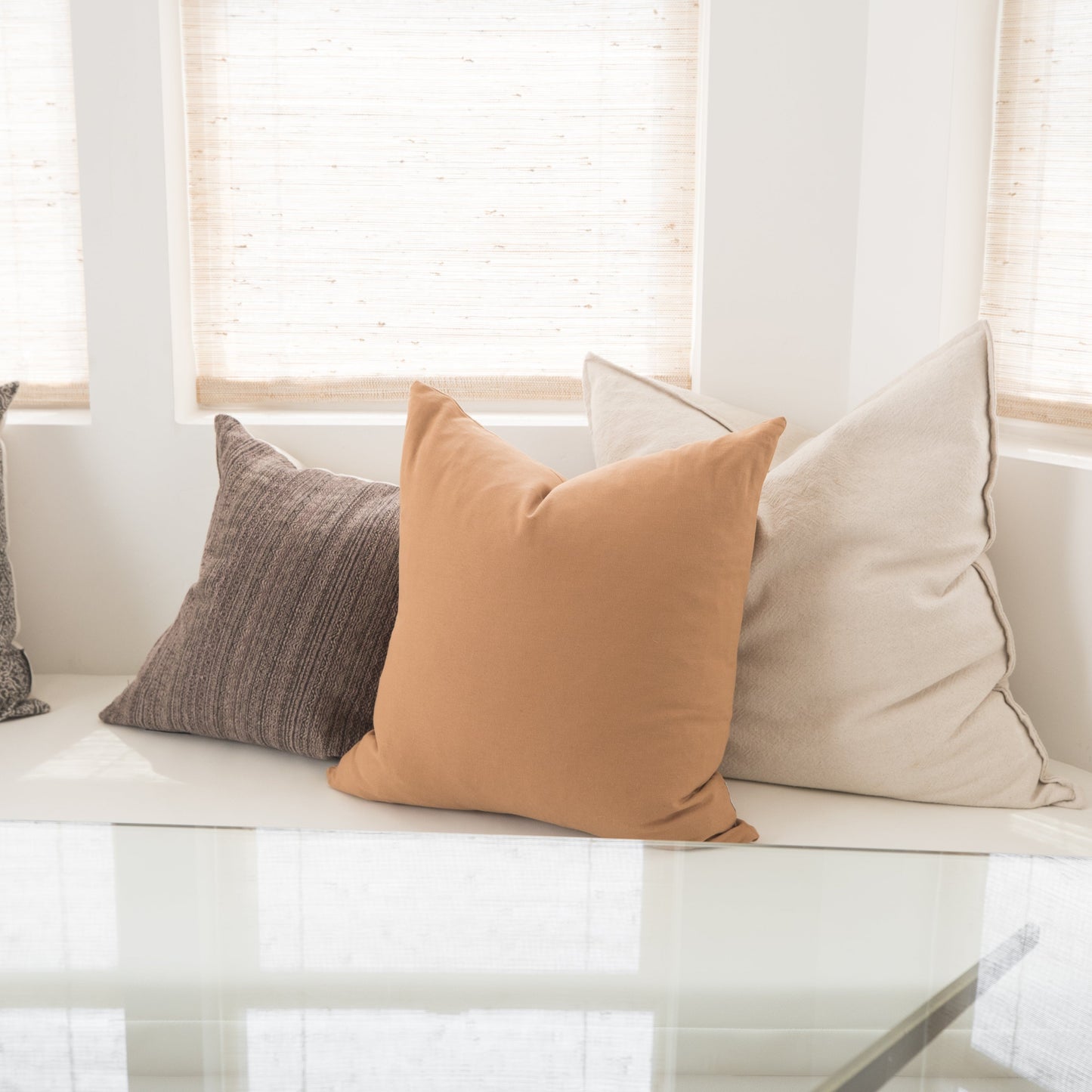 Three throw pillows in gray, terracotta, and cream arranged on a white bench by windows.