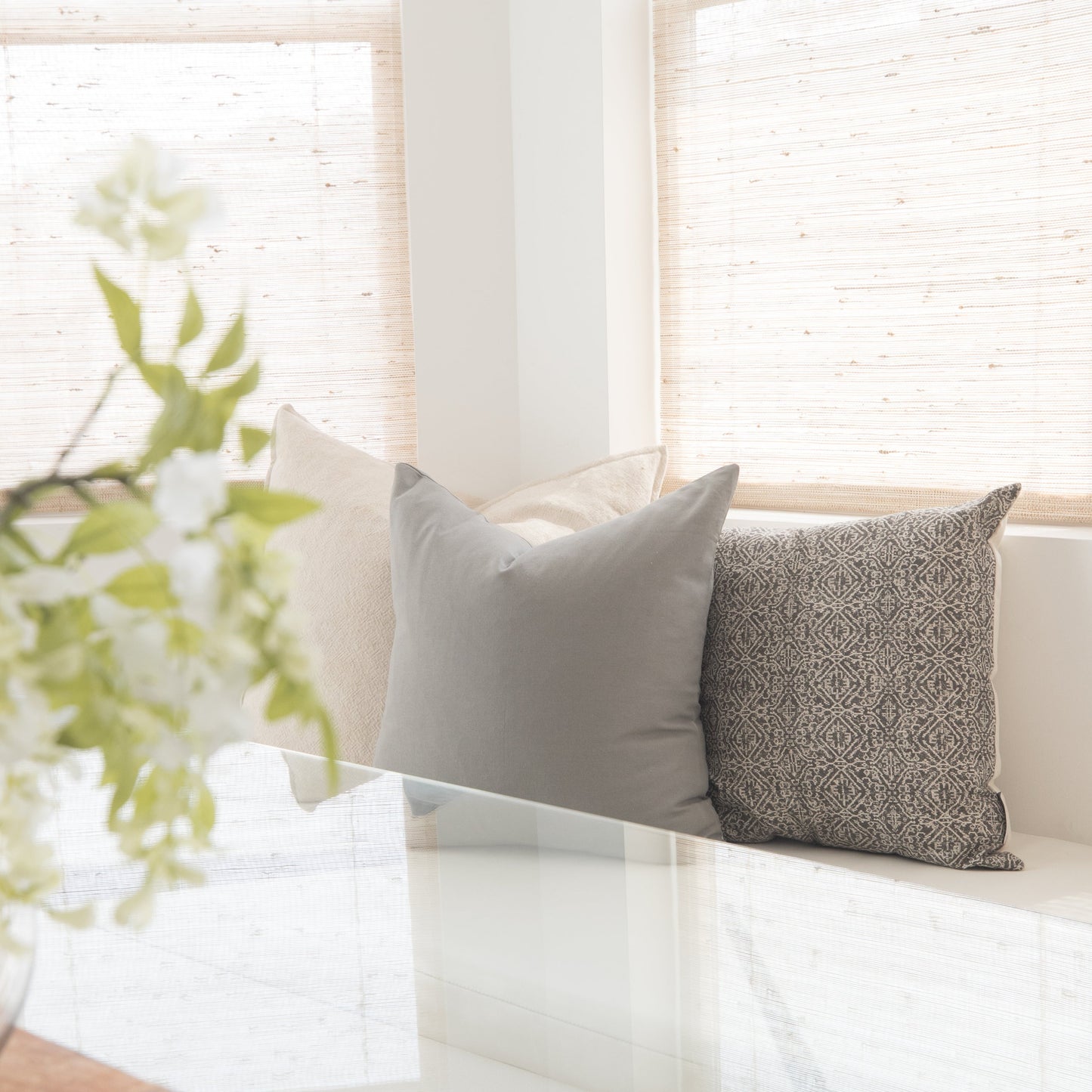 Two throw pillows in gray and patterned fabric displayed on a white surface near a window with green plant foliage.
