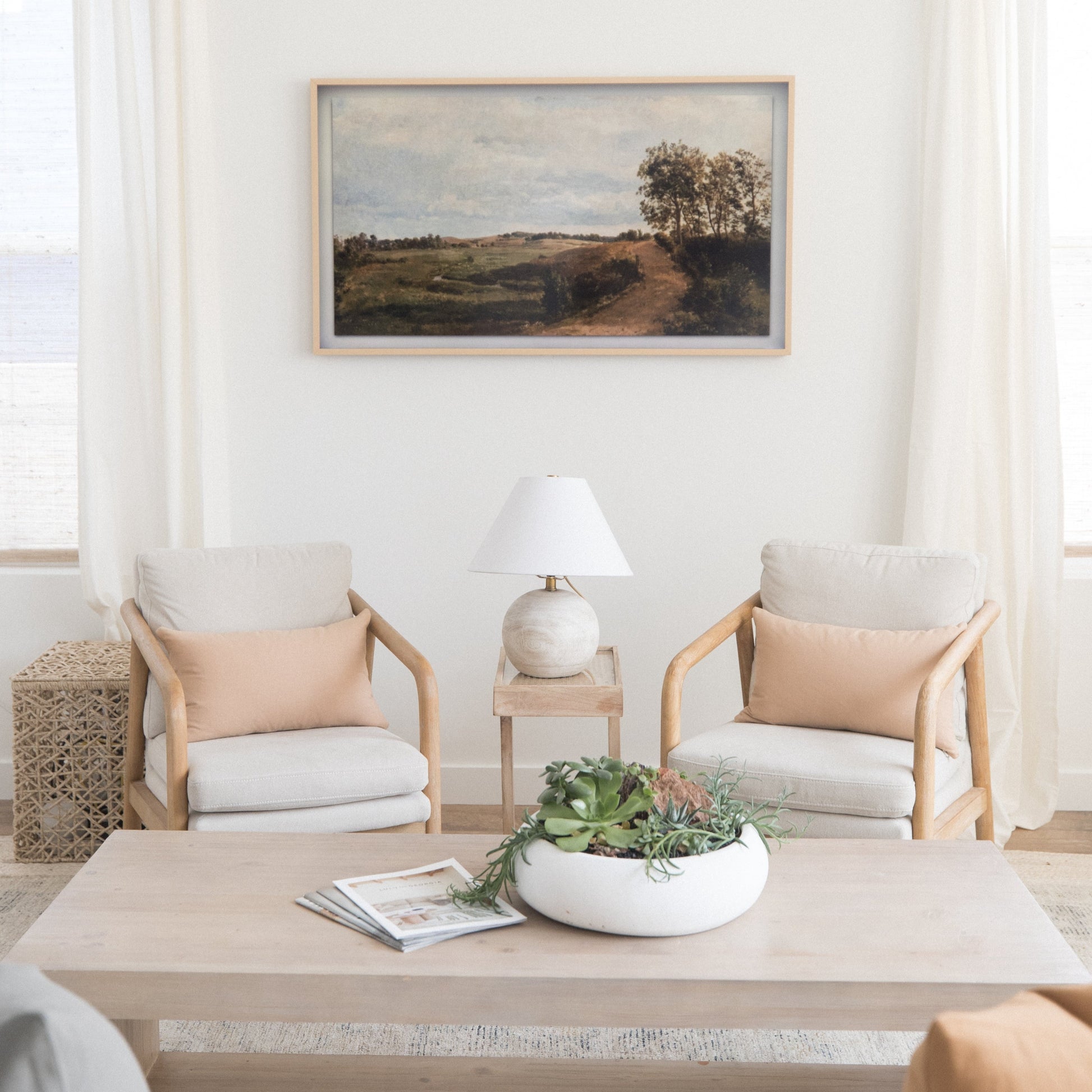 Framed landscape painting with natural wood frame hanging above two upholstered chairs in neutral tones.