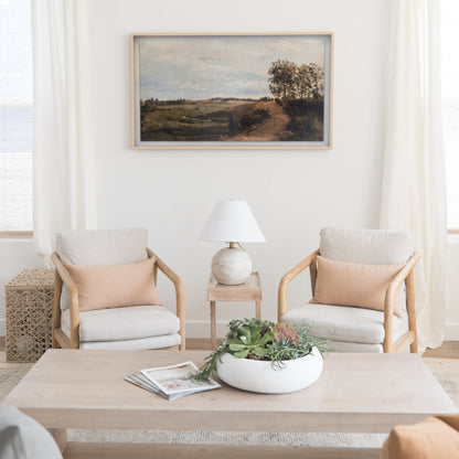Framed landscape painting with natural wood frame hanging above two upholstered chairs in neutral tones.
