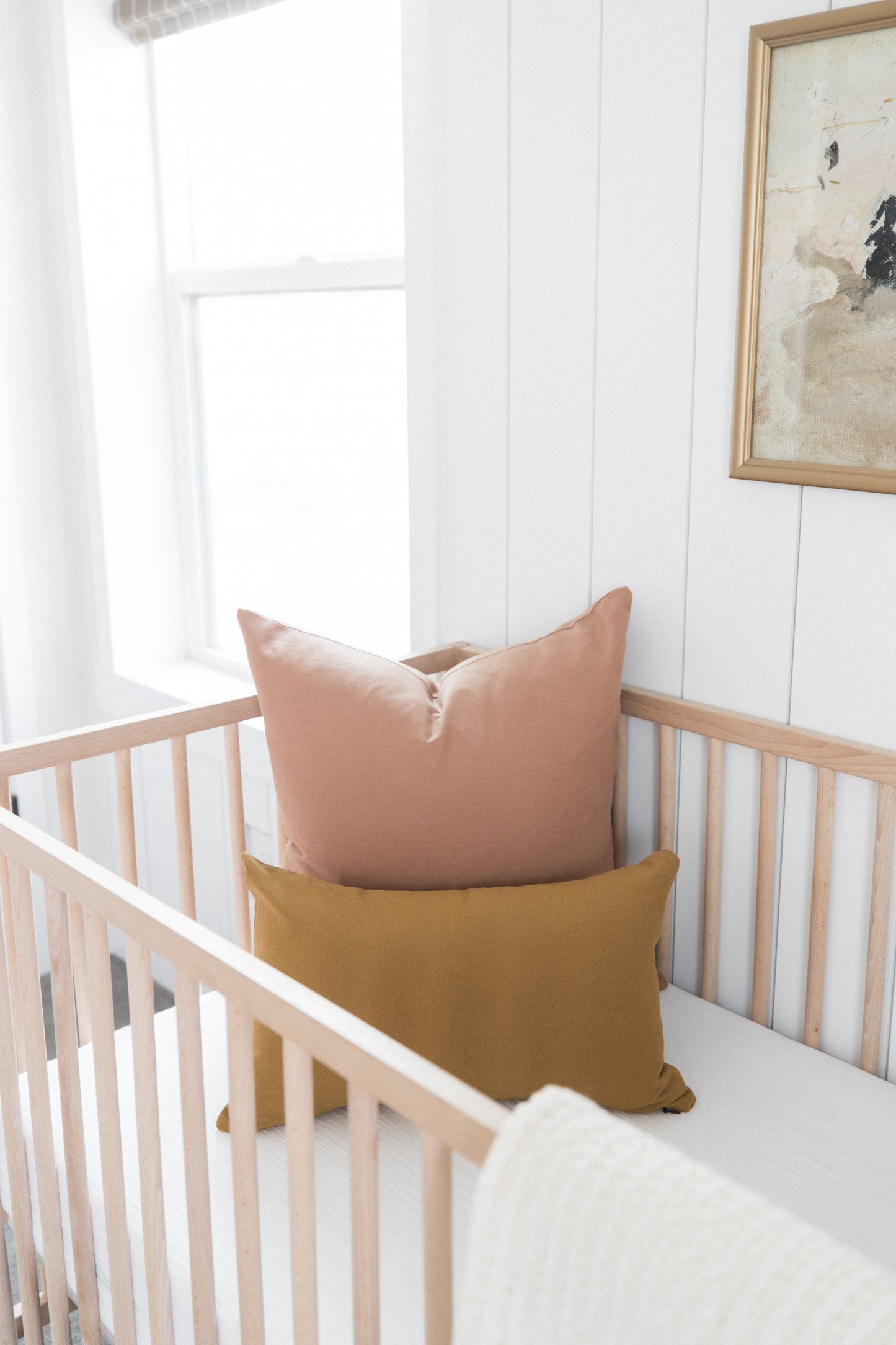 Dusty rose and mustard yellow pillows arranged in a natural wood crib with white bedding.
