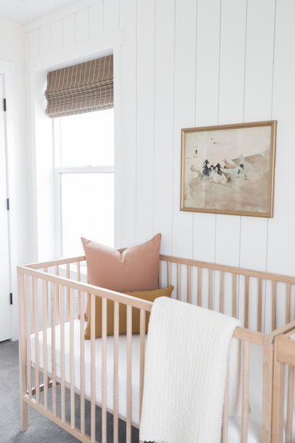 Dusty rose pillow displayed in wooden crib with white bedding in nursery room.