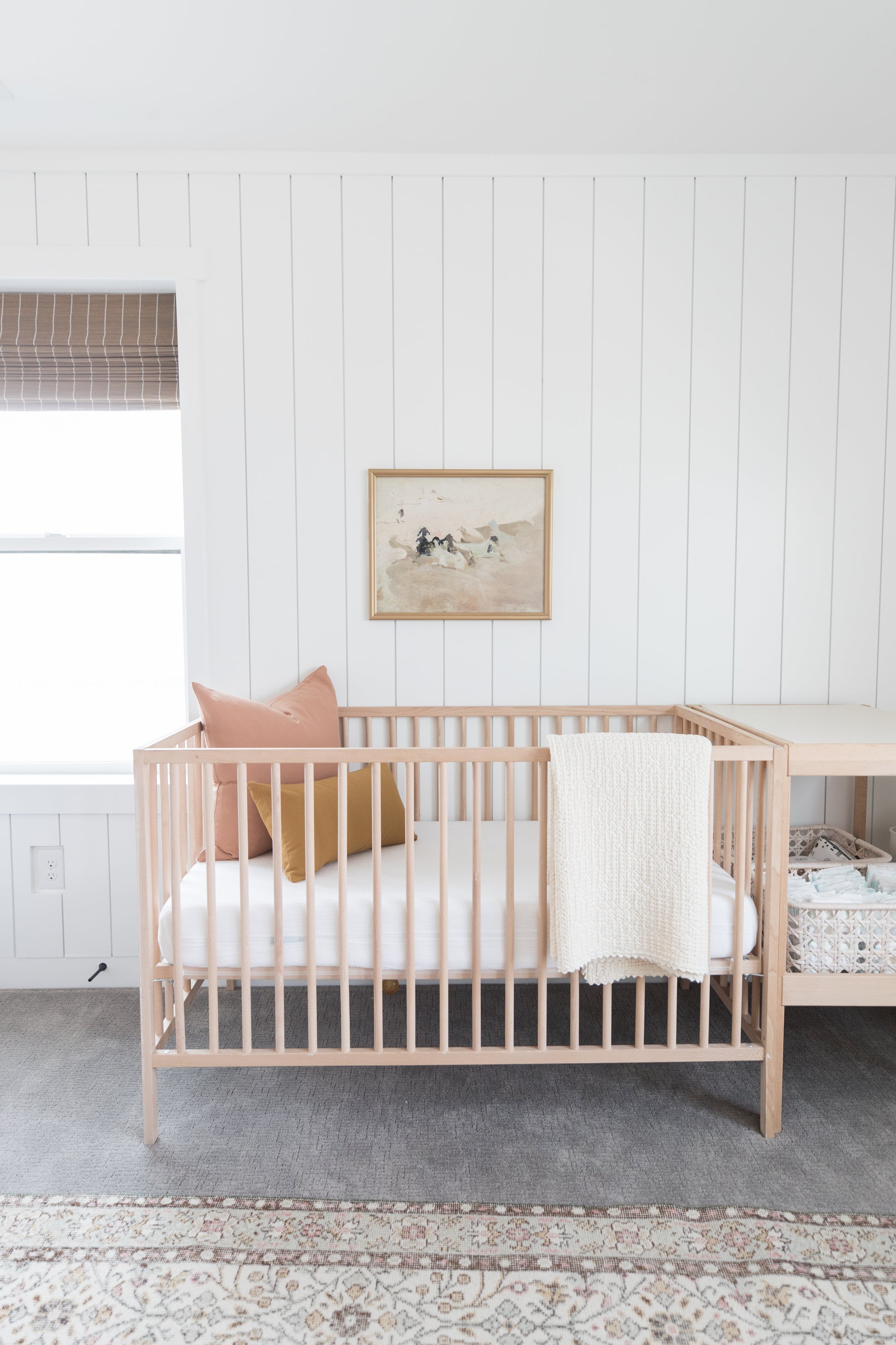 Dusty rose throw pillow displayed on wooden crib in nursery with white bedding and framed beach artwork.