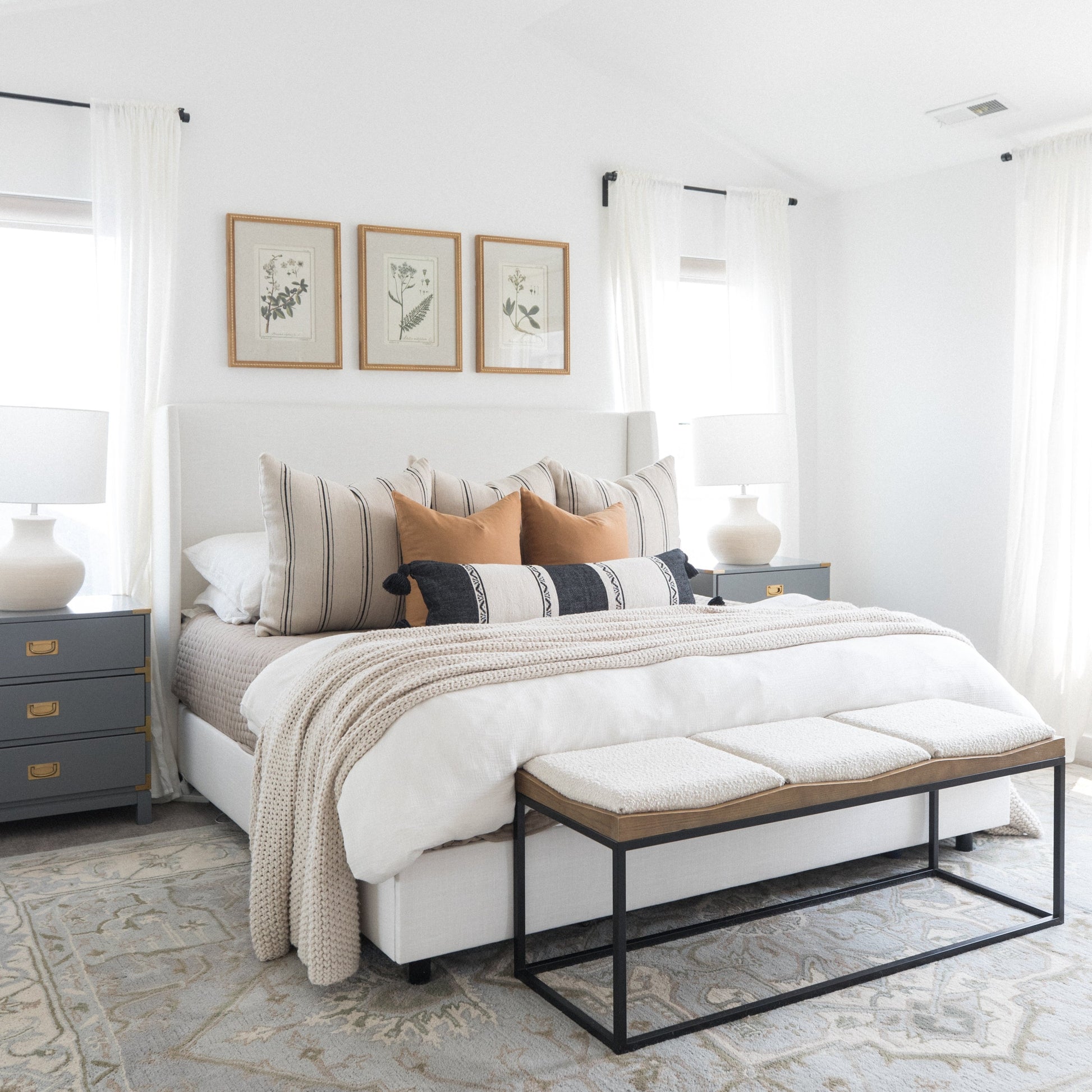 Bedroom with bed, pillows, bench, nightstand, lamp, and framed botanical prints on wall.