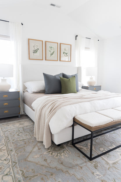 Bedroom with bed dressed in white bedding and olive green pillow, flanked by nightstands with lamps and three botanical print