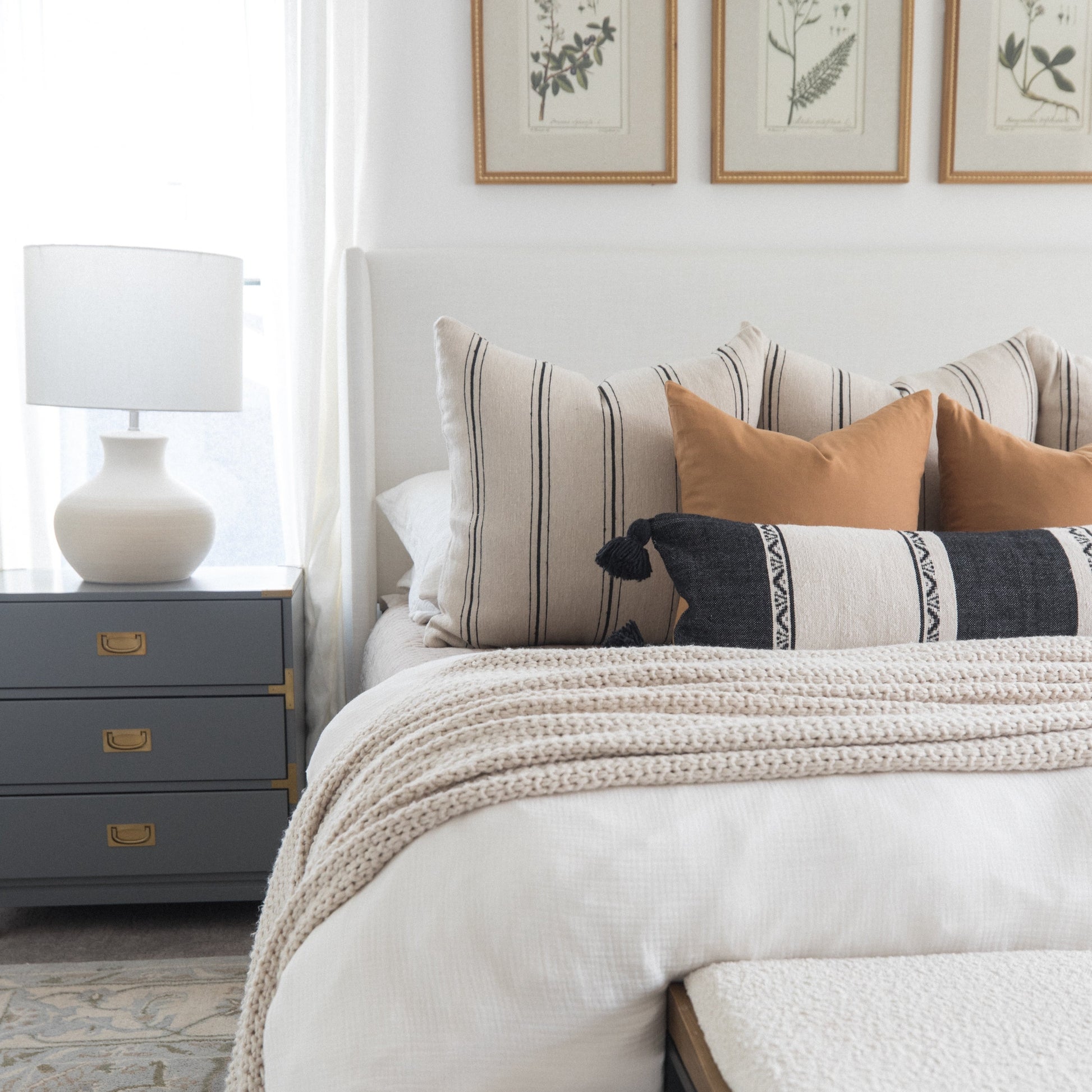 Styled bedroom scene with bed pillows, throw blanket, nightstand lamp, and framed botanical prints on wall.
