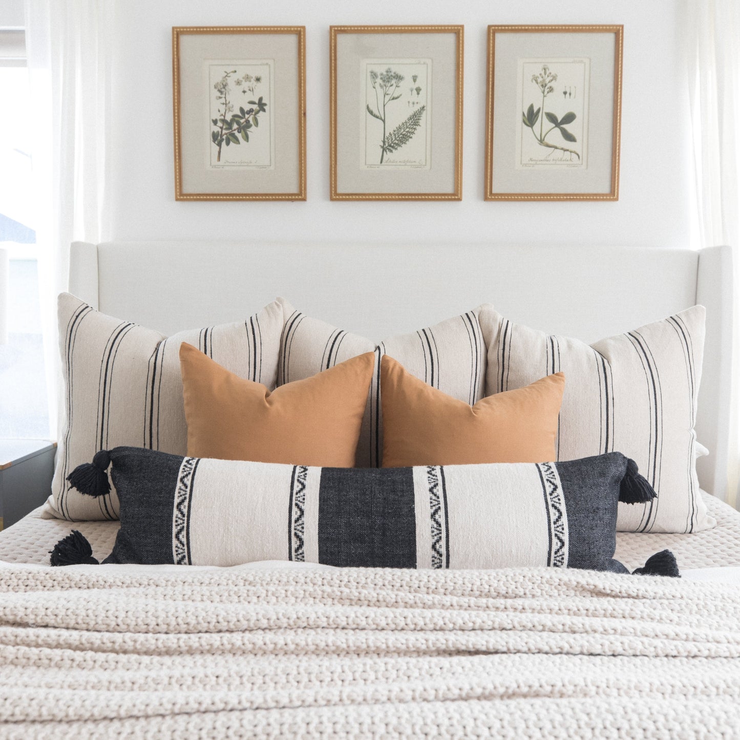 Bed styled with striped and solid terracotta pillows, patterned lumbar pillow, and three botanical framed prints above headbo