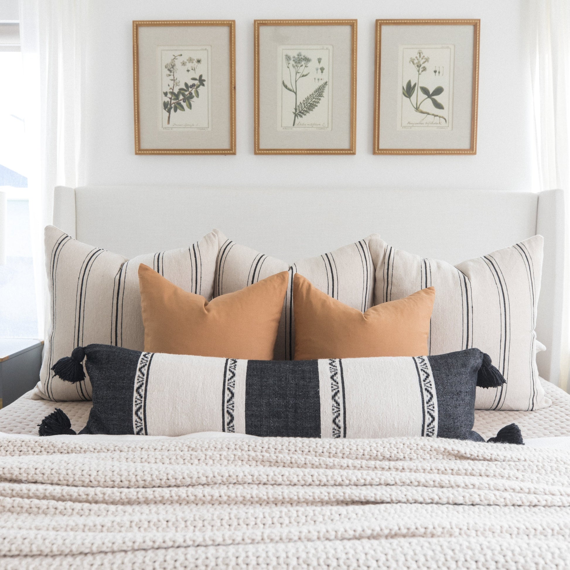 Bed styled with striped and solid terracotta pillows, patterned lumbar pillow, and three botanical framed prints above headbo