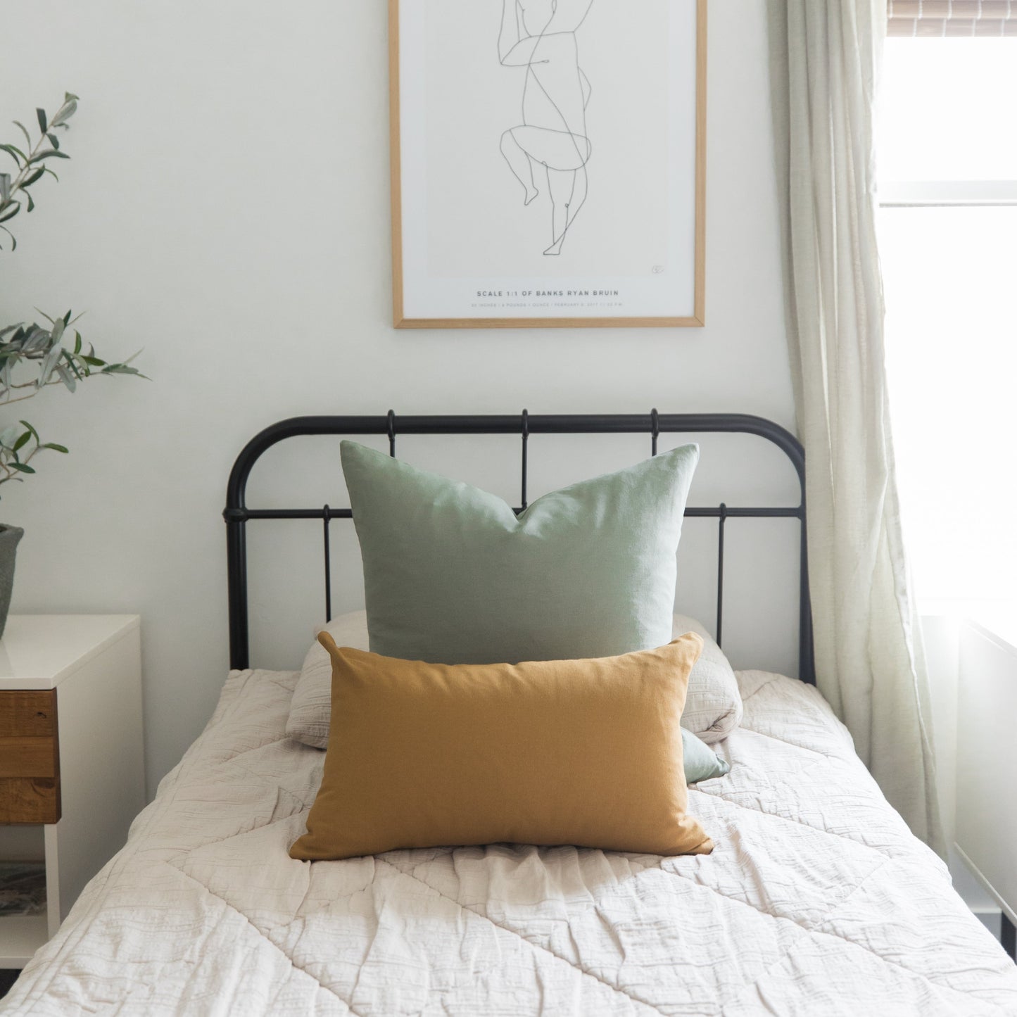 Sage green and mustard yellow pillows on a bed with black metal headboard and cream bedding.