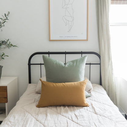 Sage green and mustard yellow pillows on a bed with black metal headboard and cream bedding.