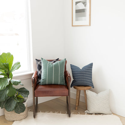 Four decorative pillows in various patterns displayed on a chair and wooden stool in a bright room.