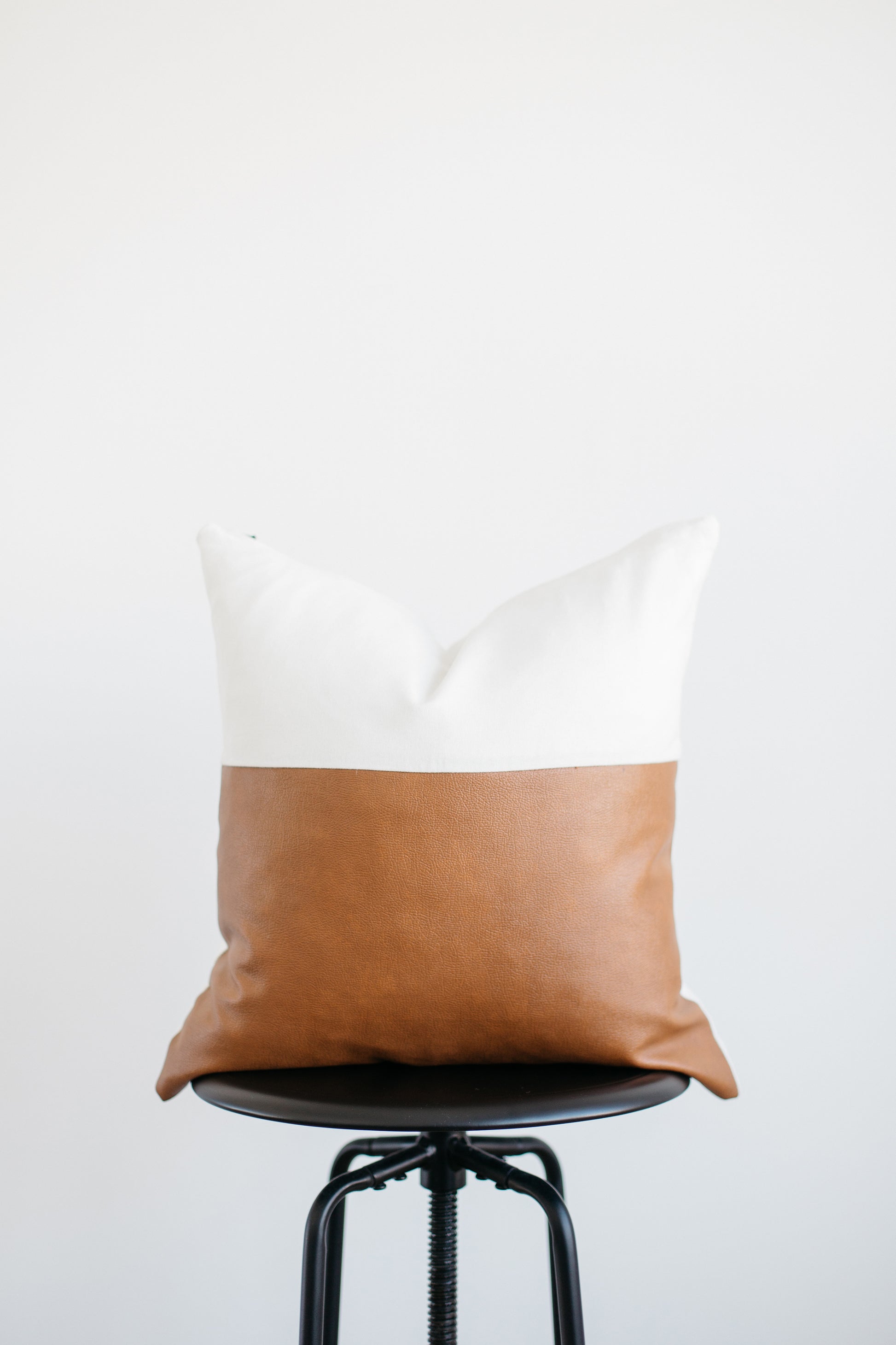 Rectangular pillow with white linen top half and tan leather bottom half, displayed on a black swivel stool.