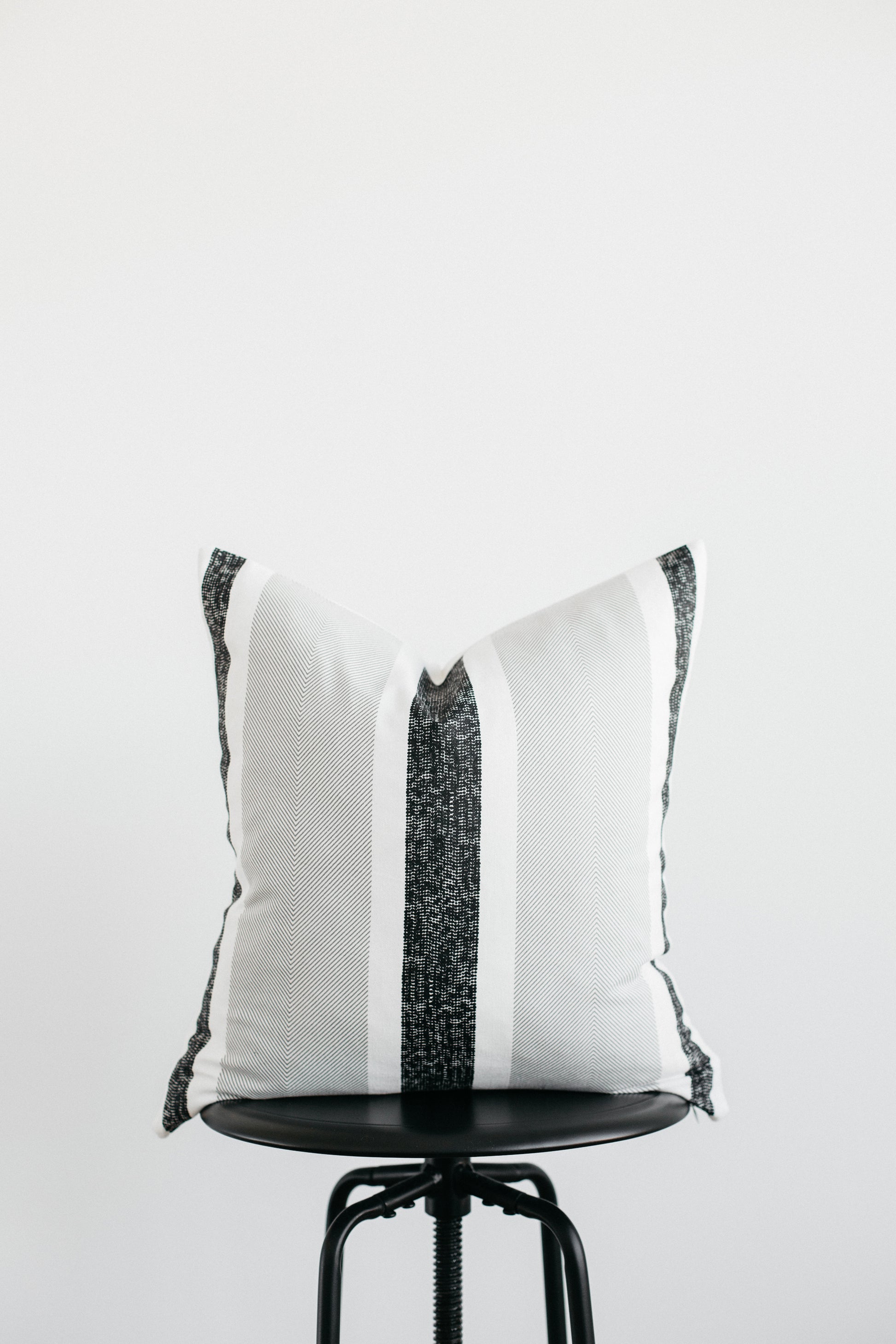 White square pillow with black vertical stripe and tassel corners displayed on black round stool.