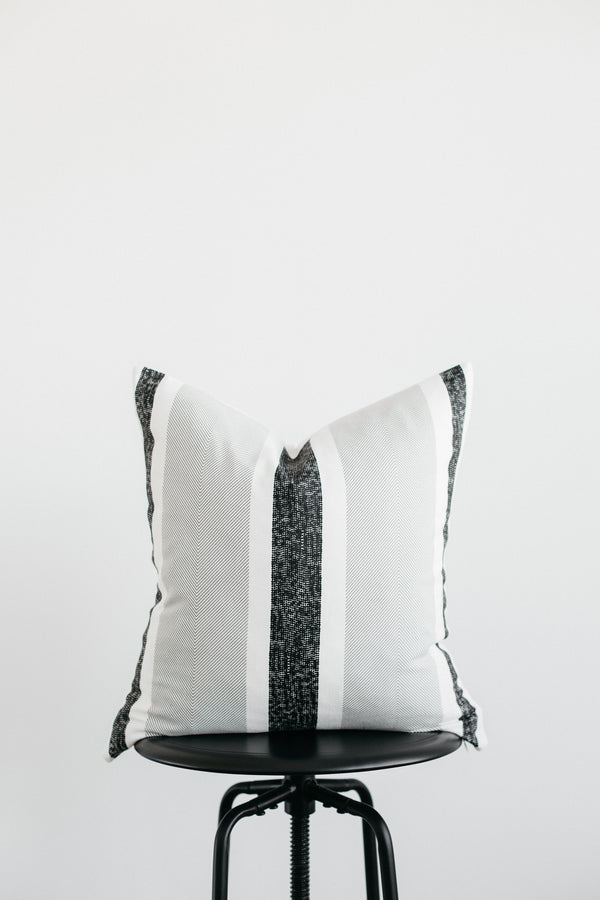 White square pillow with black vertical stripe and tassel corners displayed on black round stool.
