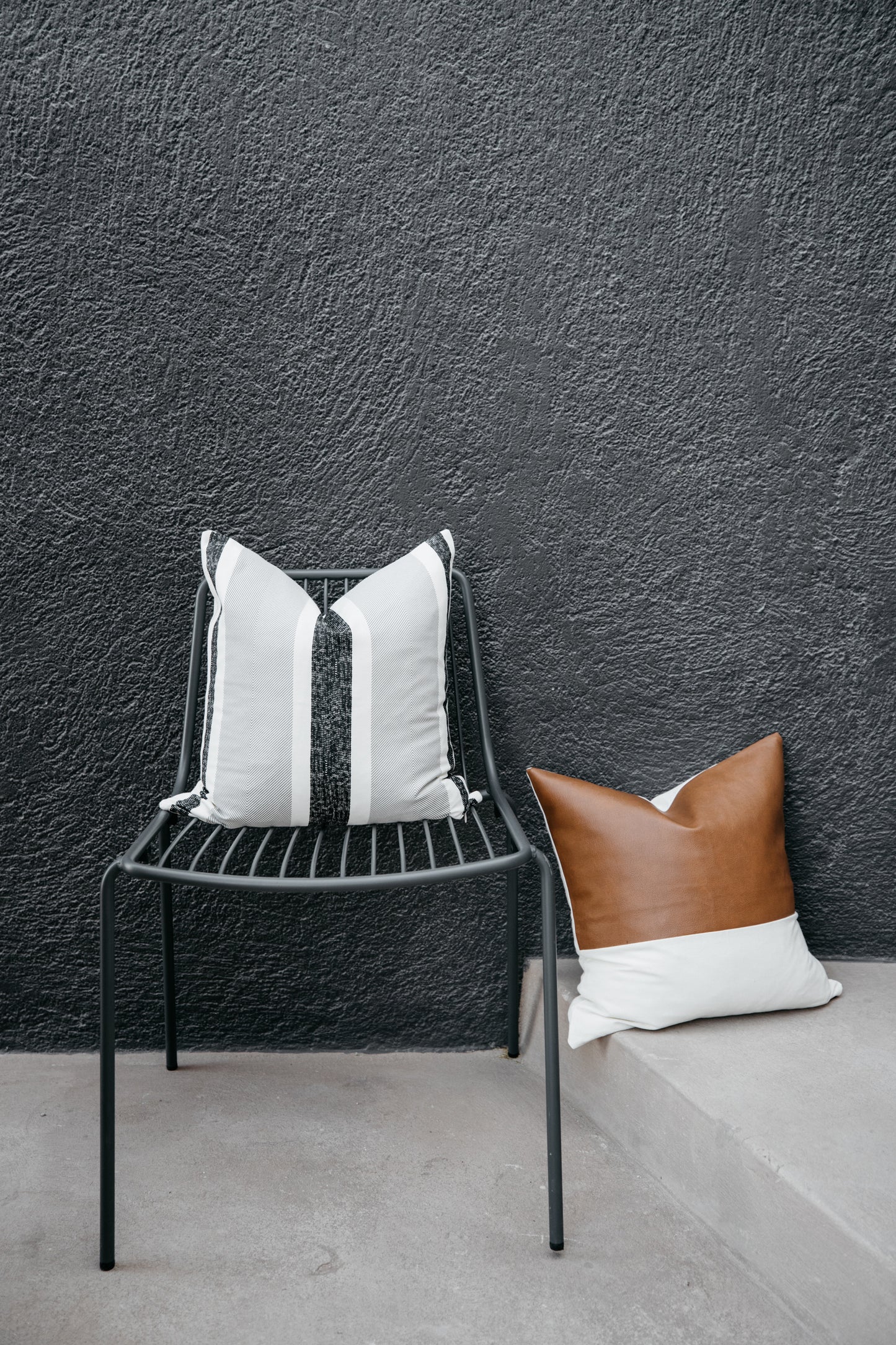 Black metal chair with white and gray striped pillow, brown leather pillow on concrete ledge against textured gray wall.