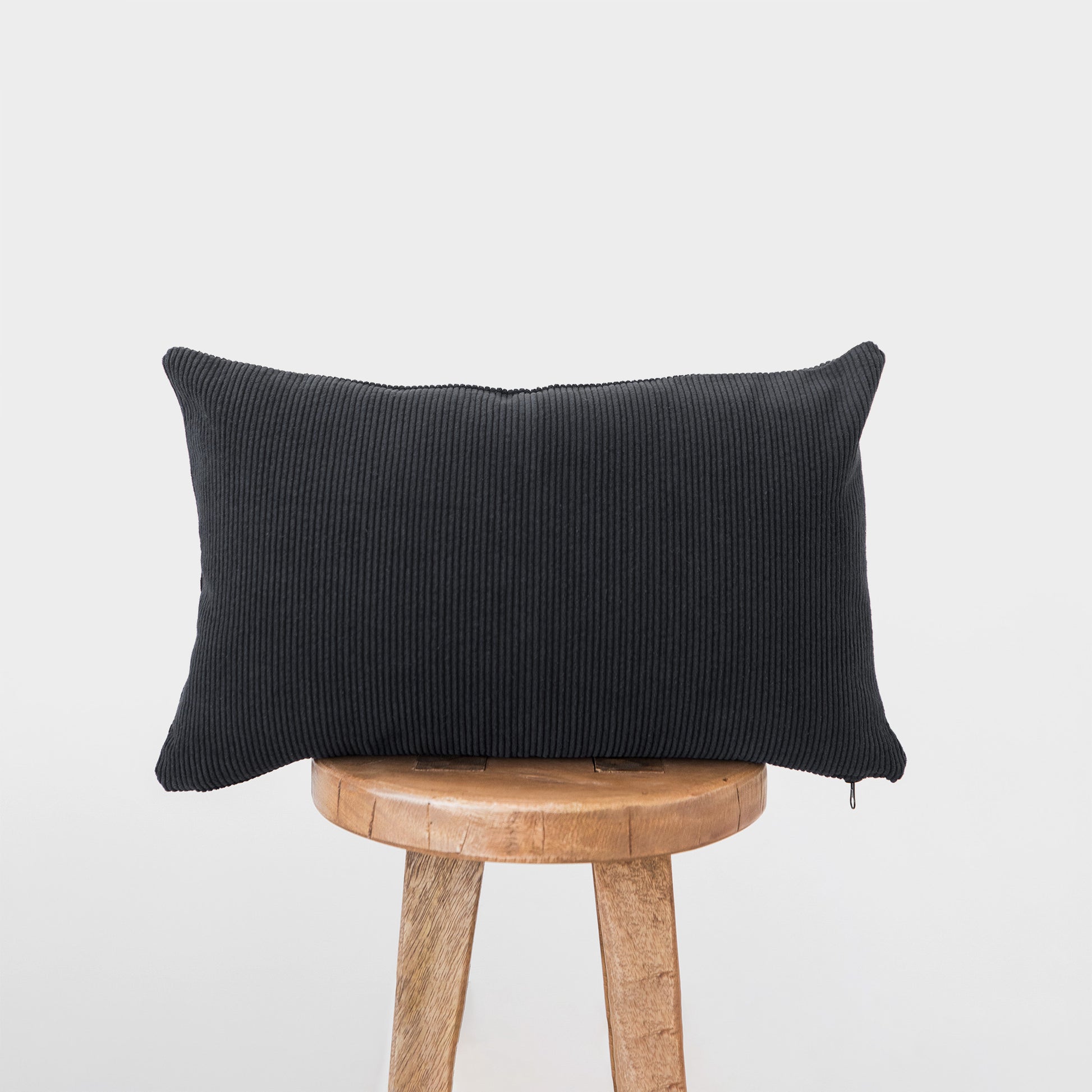 Dark gray corduroy lumbar pillow with ribbed texture displayed on a wooden stool.