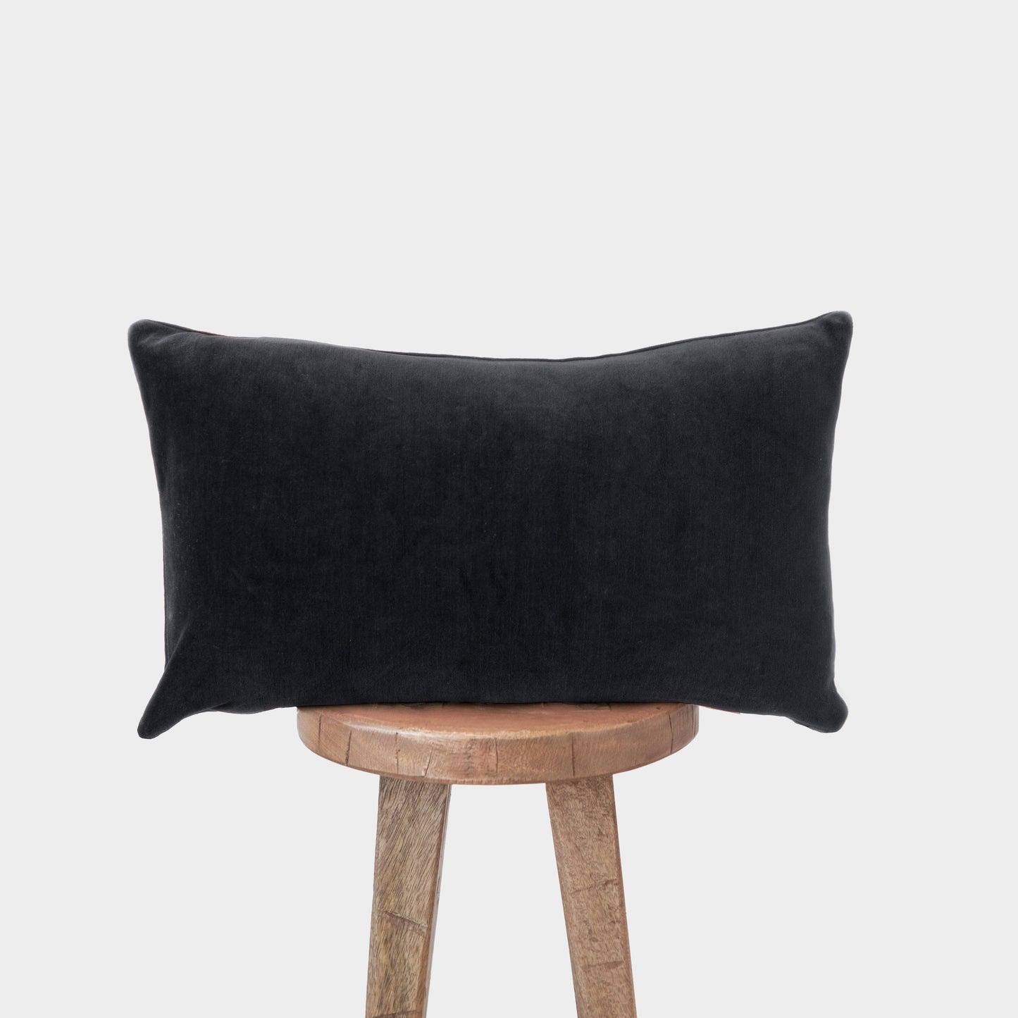 Black velvet lumbar pillow with plump rectangular shape displayed on wooden stool.