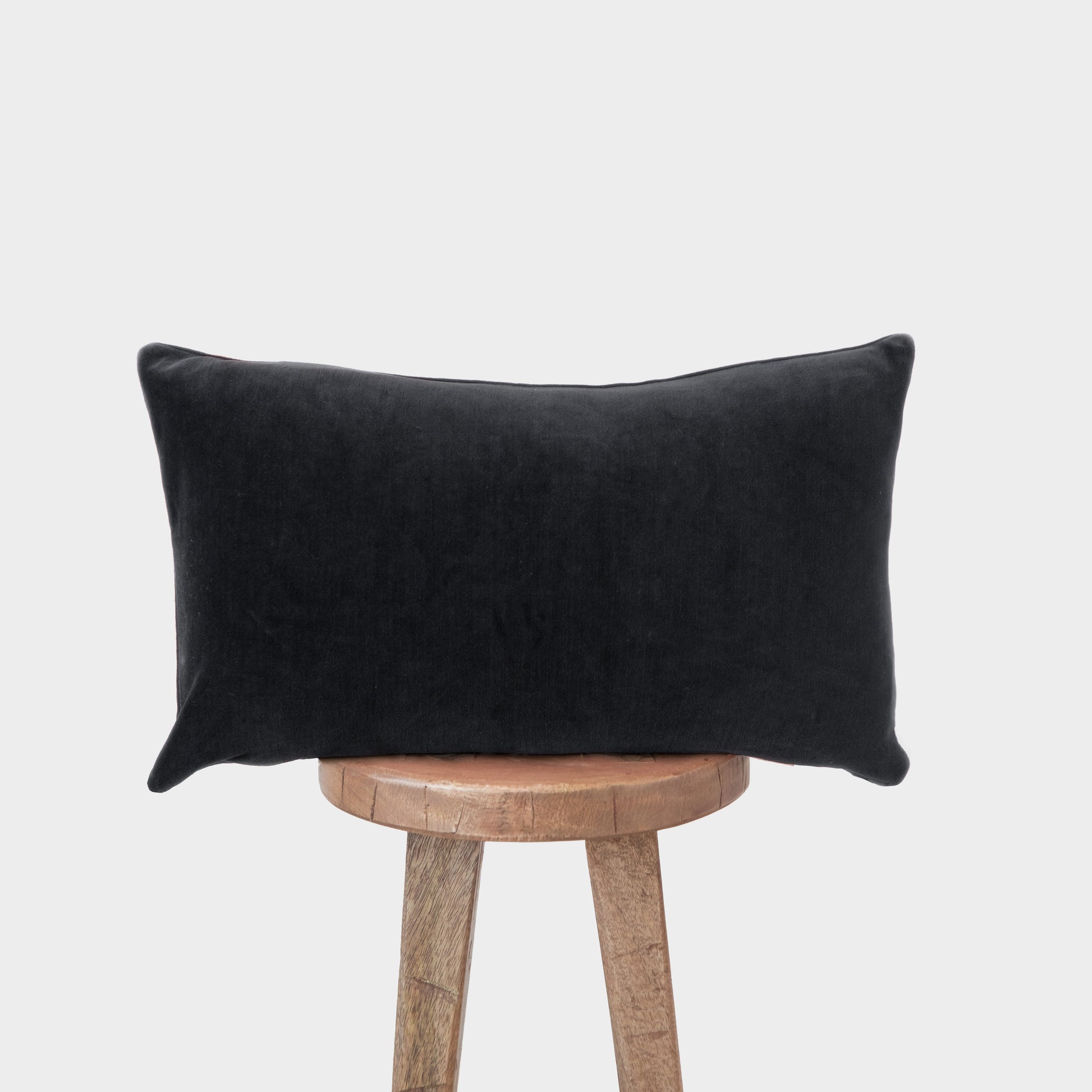 Black velvet lumbar pillow with plump rectangular shape displayed on wooden stool.