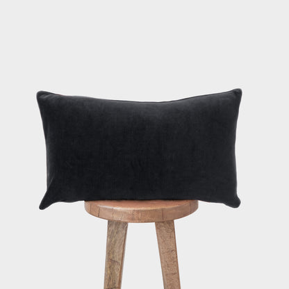 Black velvet lumbar pillow with plump rectangular shape displayed on wooden stool.