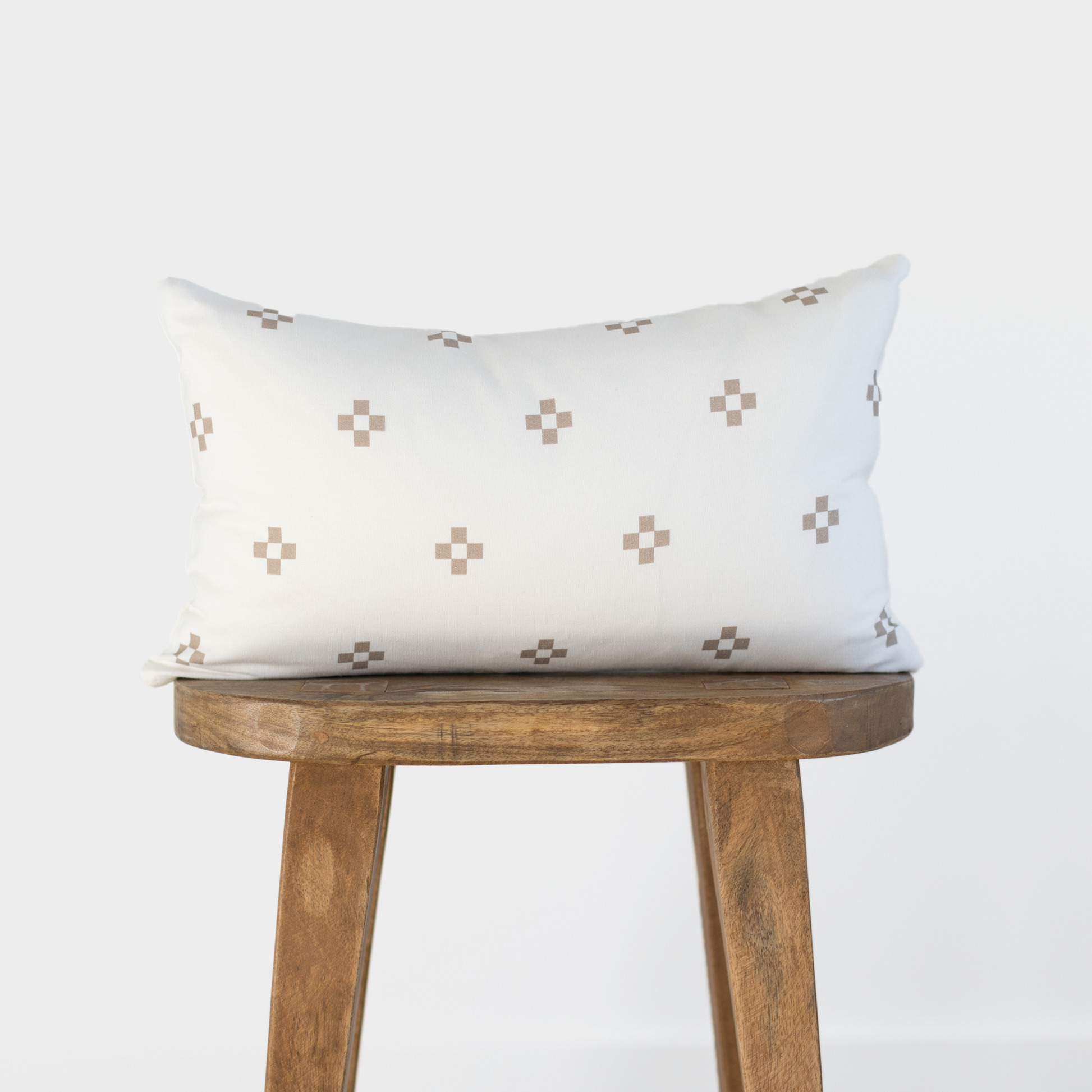 Cream lumbar pillow with tan square dot pattern displayed on wooden stool.