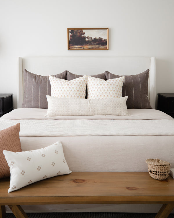 Cream lumbar pillow with small geometric floral pattern displayed on wooden bench in front of bed.