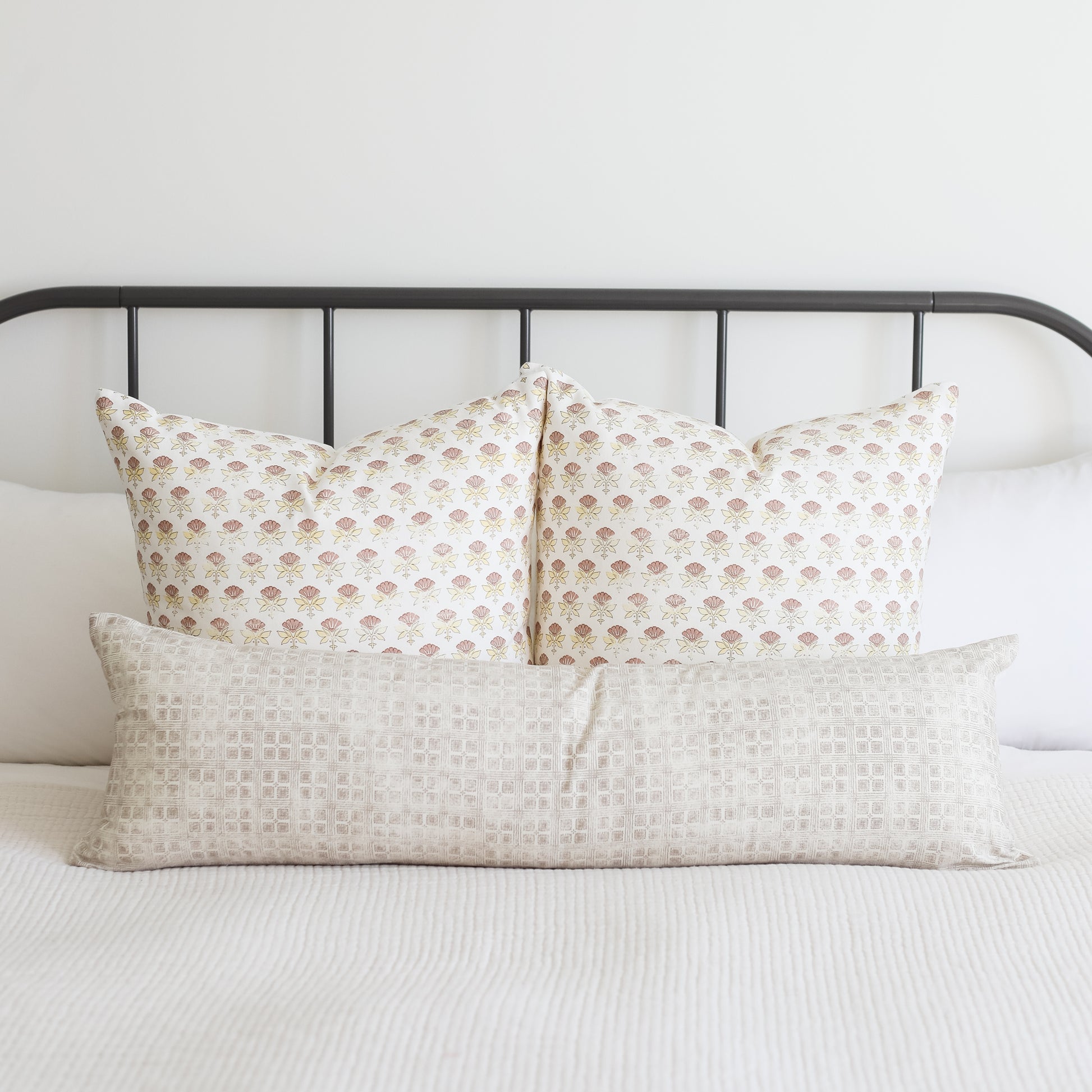 White decorative pillows with small floral pattern arranged on a metal bed frame with white bedding.