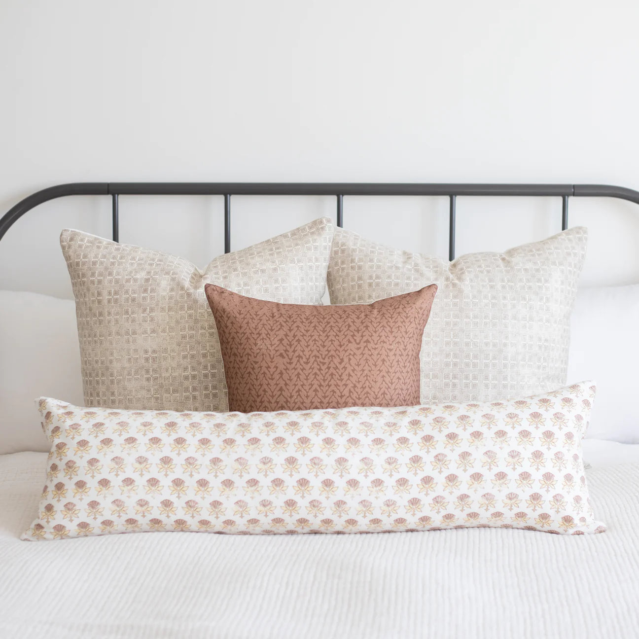 White lumbar pillow with small tan floral pattern, displayed among coordinating neutral and patterned pillows on a bed.