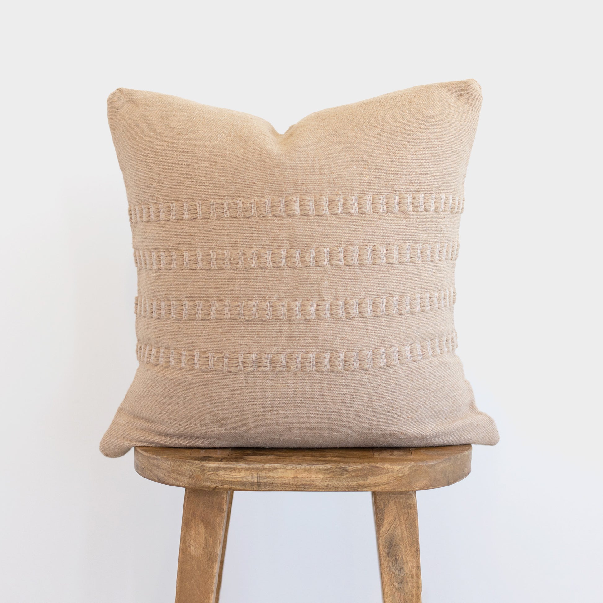Tan linen pillow with horizontal woven stripe details, displayed on a wooden stool.