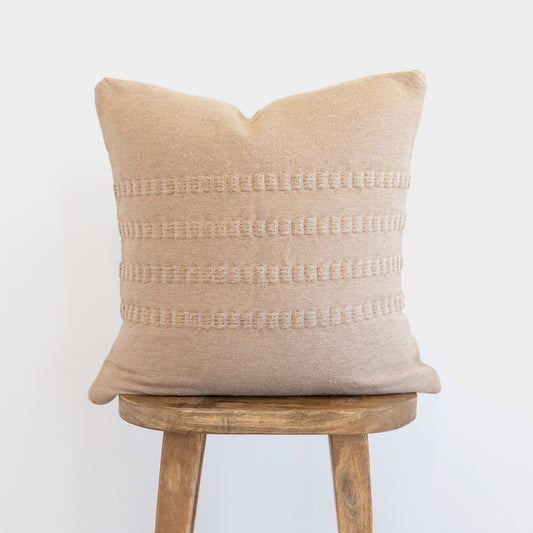 Tan linen pillow with horizontal woven stripe details, displayed on a wooden stool.