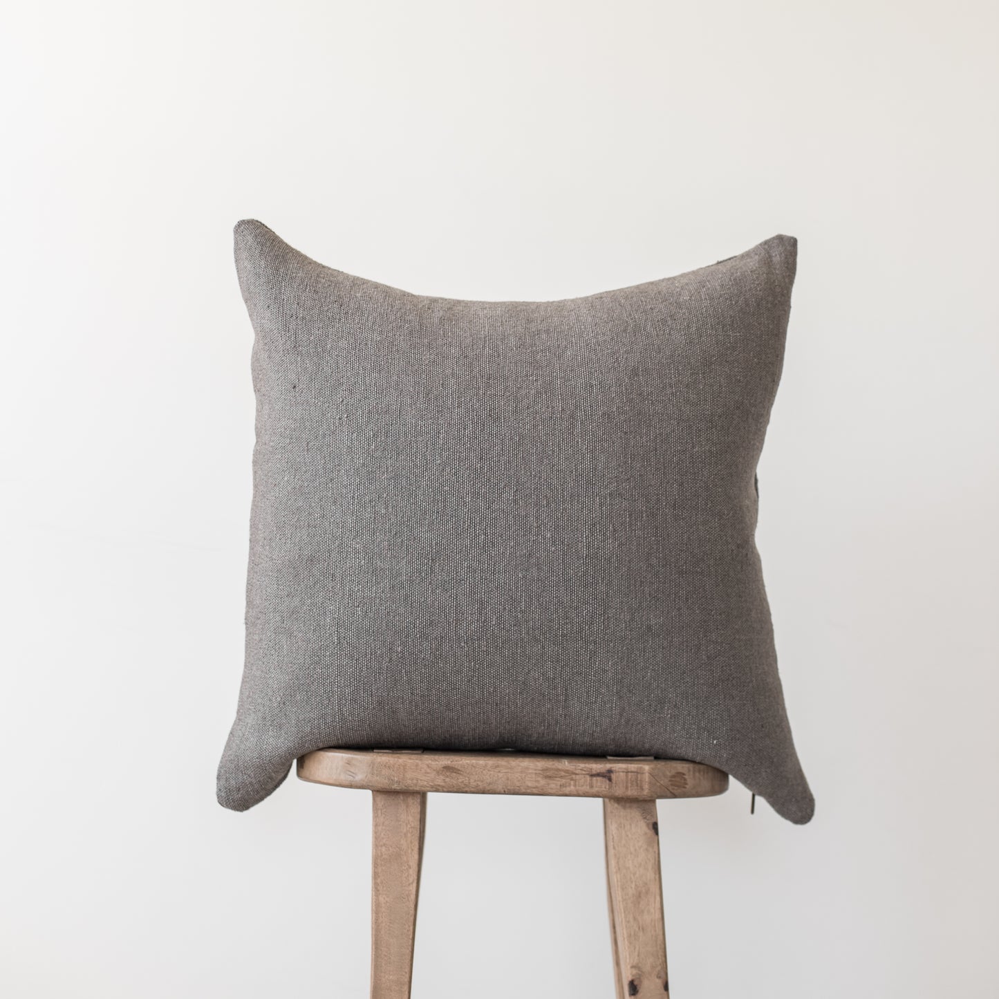 Gray linen throw pillow with knife edges displayed on wooden stool.