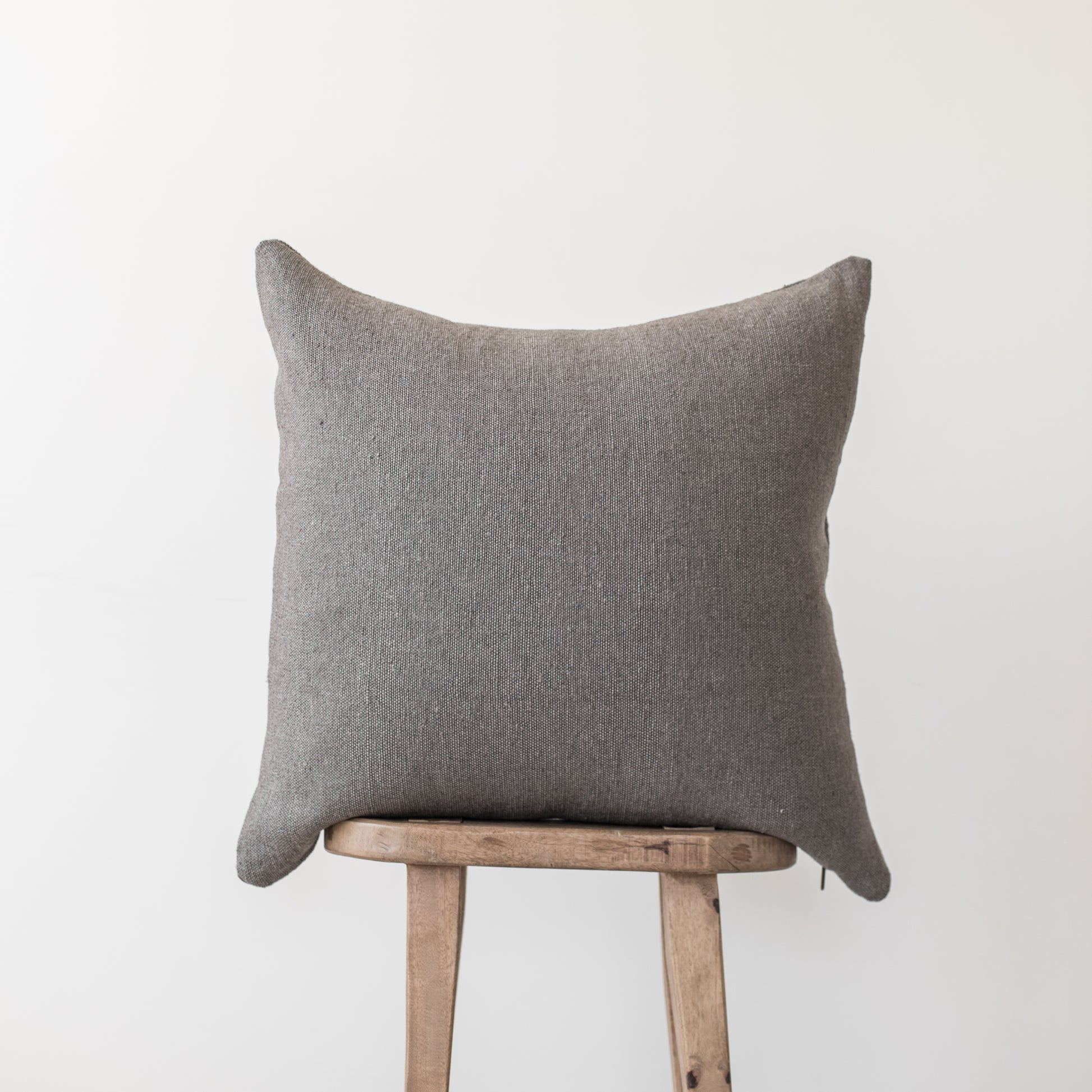 Gray linen throw pillow with knife edges displayed on wooden stool.