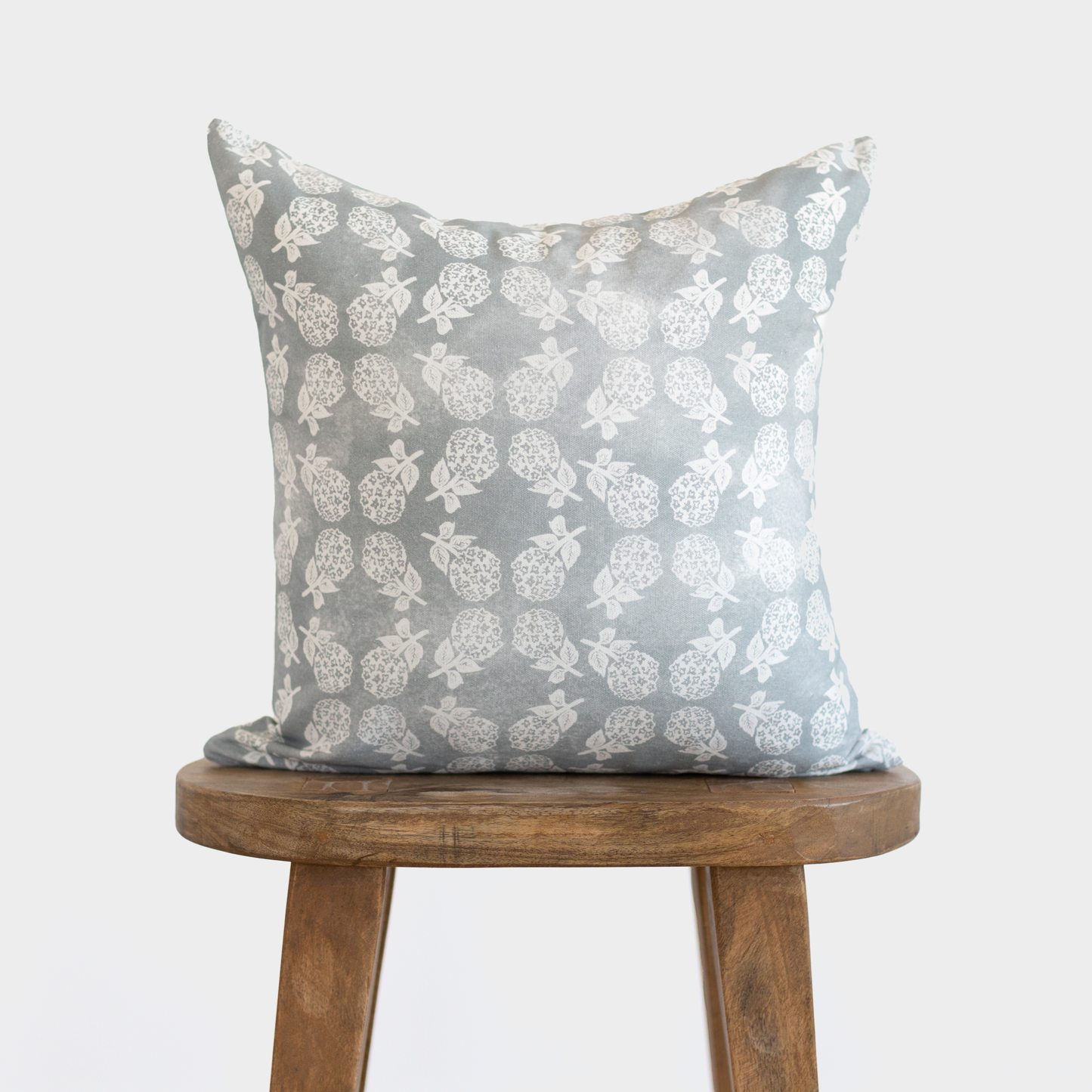 Gray linen pillow with white pineapple and leaf pattern, displayed on wooden stool.