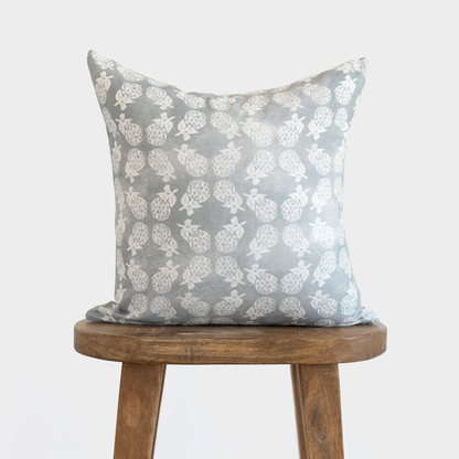 Gray linen pillow with white pineapple and leaf pattern, displayed on wooden stool.