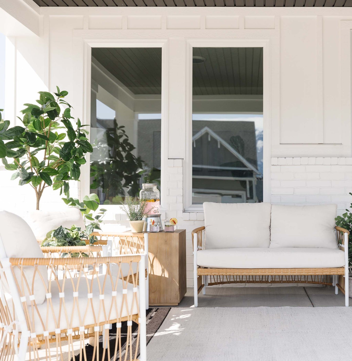 Covered porch with woven rattan furniture, white cushions, potted plants, and wooden side table.