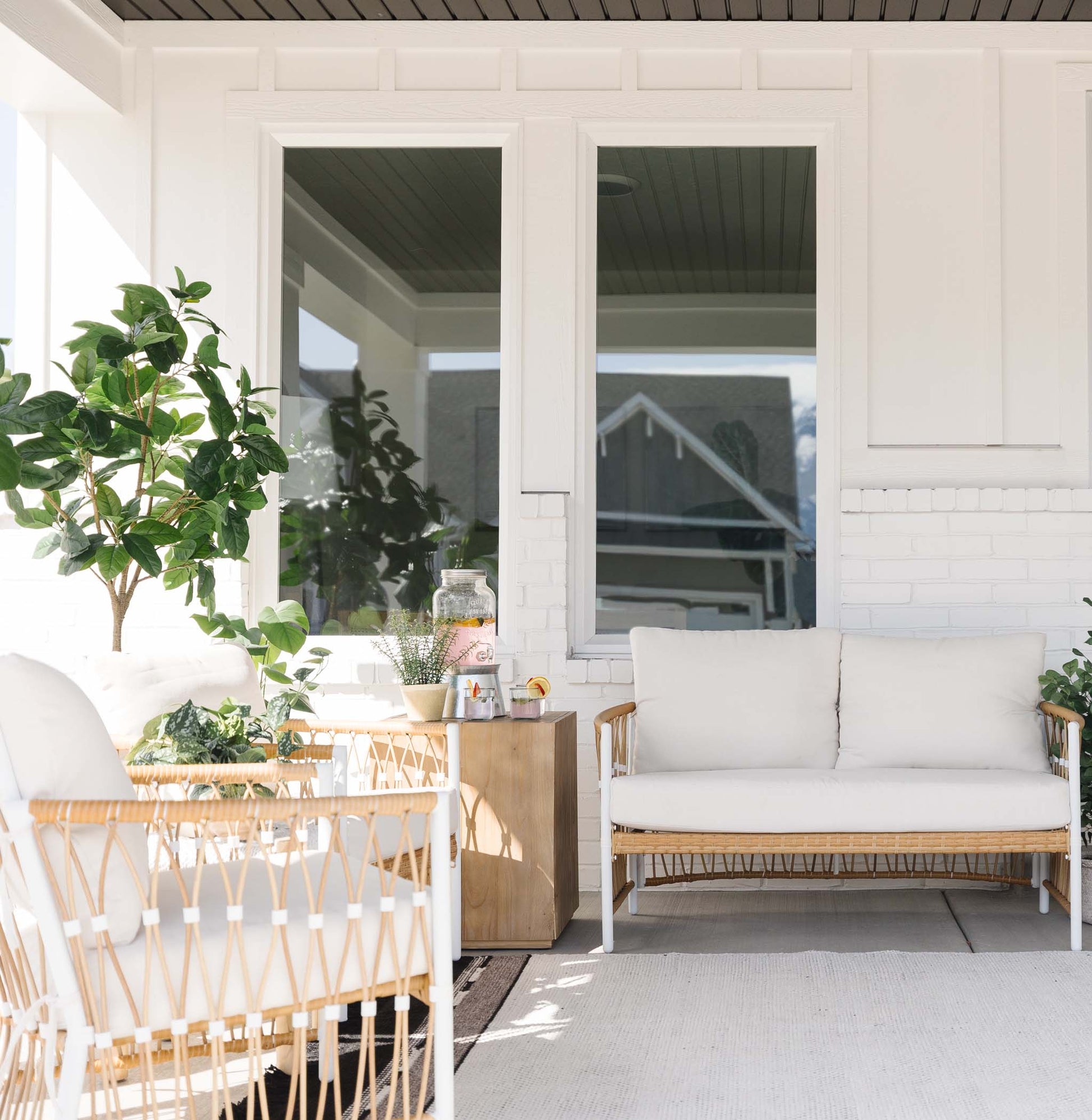 Covered porch with woven rattan furniture, white cushions, potted plants, and wooden side table.