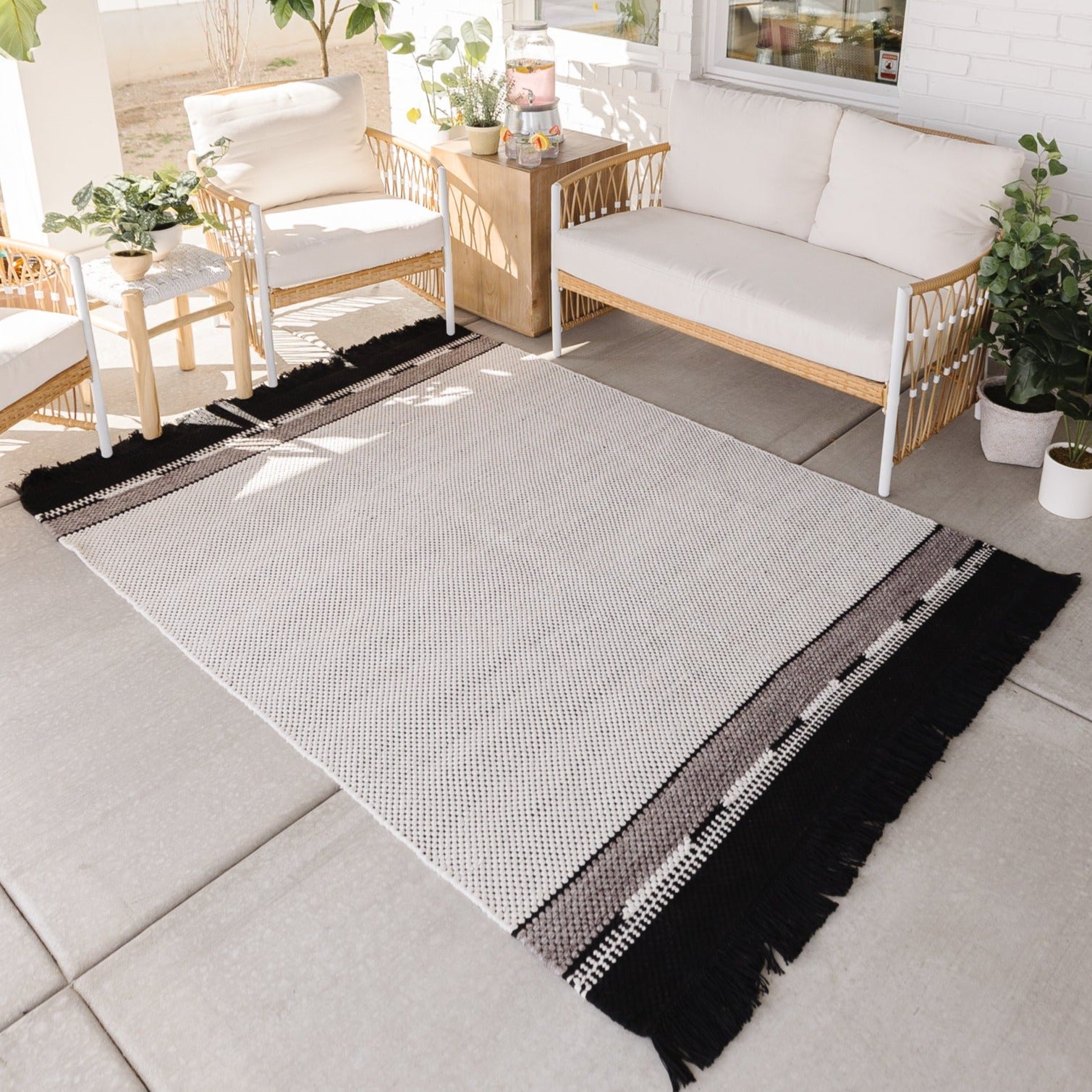Light gray textured outdoor rug with black and white striped border and fringe, displayed on patio with wooden furniture.