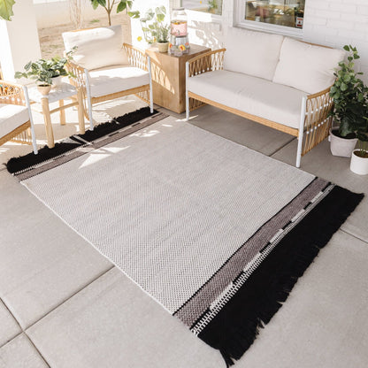 Light gray textured outdoor rug with black and white striped border and fringe, displayed on patio with wooden furniture.