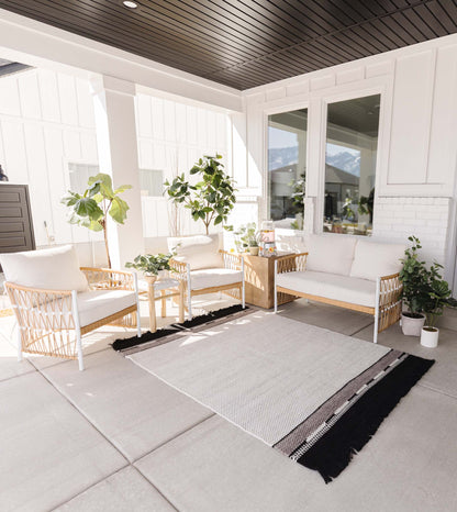 Light gray and black striped outdoor rug with fringe edges displayed on a covered patio with wicker furniture.