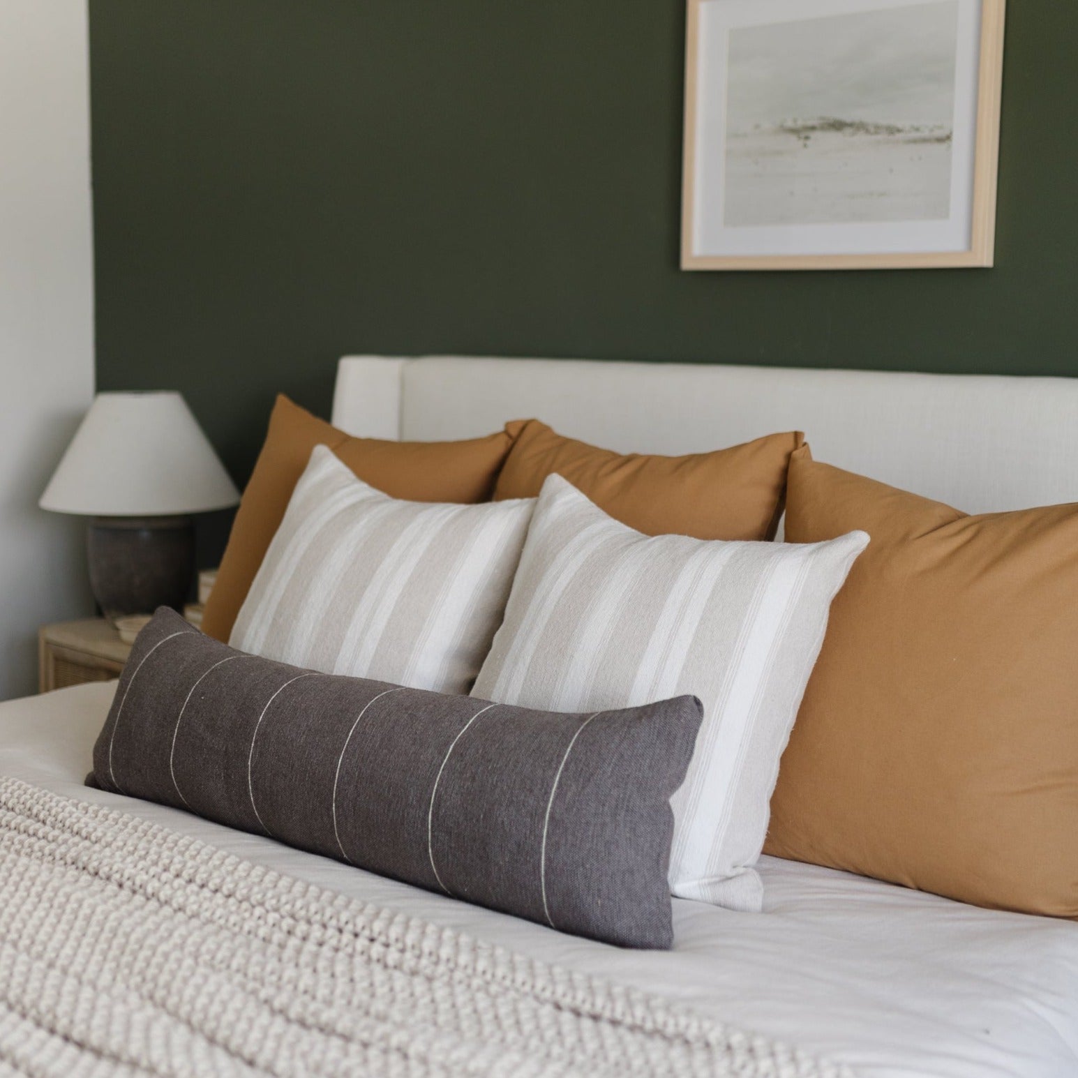 Tan lumbar pillow with vertical stitching detail displayed on bed with striped and solid pillows.