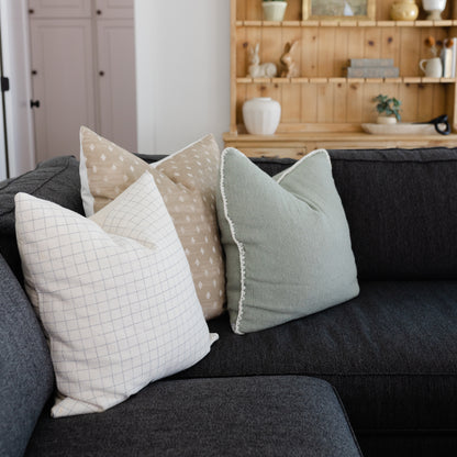 Three decorative pillows with grid and dot patterns in cream, tan, and sage green on a dark gray sofa.