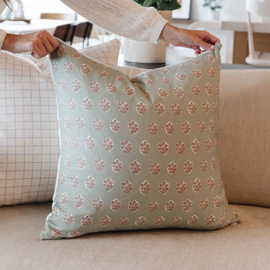Sage green throw pillow with repeating rose gold damask pattern, held by hands over furniture.