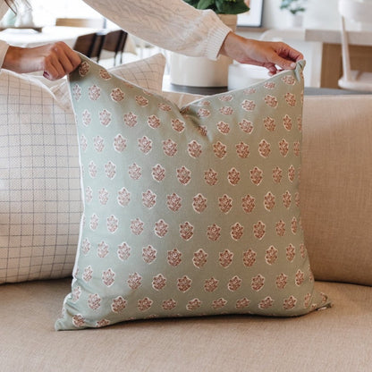 Sage green throw pillow with repeating rose gold damask pattern, held by hands over furniture.