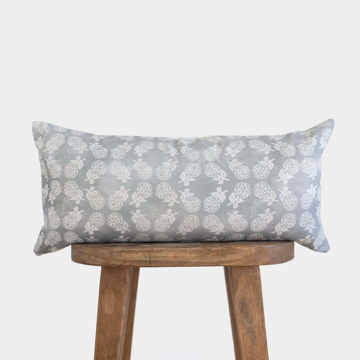 Gray rectangular lumbar pillow with cream pineapple and leaf pattern, displayed on wooden stool.