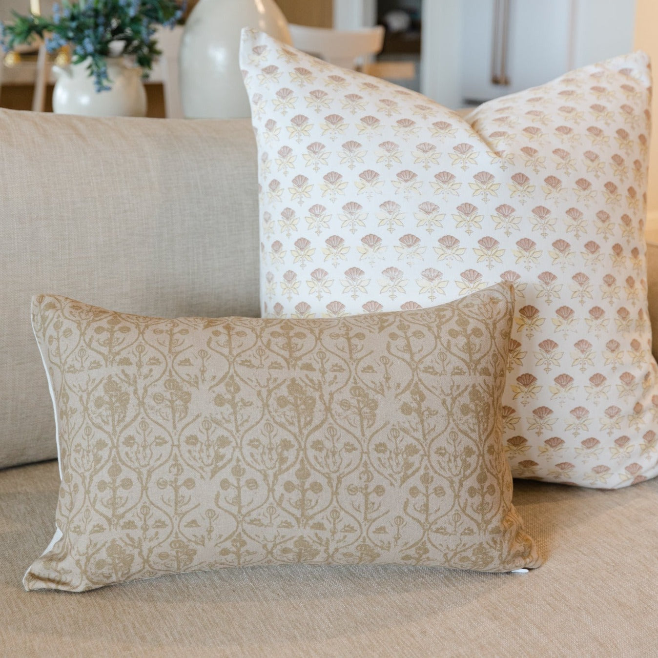 Beige and cream floral patterned throw pillows arranged on a light upholstered sofa with decorative vase in background.